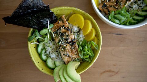 A yellow bowl filled with rice, sliced avocado, cucumber, pickled radish, and grilled fish topped with herbs, seaweed, and green onions—a vibrant Salmon Rice Bowl Recipe. Another similar bowl is partially visible in the background on a wooden surface.