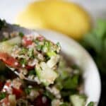 A close-up of a spoonful of taboule salad with chopped parsley, tomatoes, cucumbers, and bulgur. A bowl of taboule, fresh parsley, and a lemon are visible in the blurred background.
