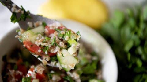 A close-up of a spoonful of taboule salad with chopped parsley, tomatoes, cucumbers, and bulgur. A bowl of taboule, fresh parsley, and a lemon are visible in the blurred background.