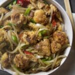 A bowl of salt and pepper chicken features crispy pieces stir-fried with sliced onions, green onions, and red chili peppers, placed on a white napkin with chopsticks beside it.