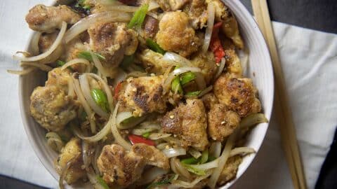 A bowl of salt and pepper chicken features crispy pieces stir-fried with sliced onions, green onions, and red chili peppers, placed on a white napkin with chopsticks beside it.