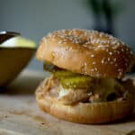 A close-up of an Alabama Chicken Sandwich with a sesame seed bun, shredded chicken, pickles, and sauce sits on a wooden surface next to a yellow bowl with a spoon.
