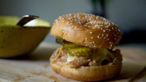 A close-up of an Alabama Chicken Sandwich with a sesame seed bun, shredded chicken, pickles, and sauce sits on a wooden surface next to a yellow bowl with a spoon.