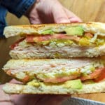 A person holds an Albuquerque Turkey Sandwich cut in half, revealing layers of sliced turkey, avocado, tomato, and possibly cheese on toasted bread, with a wooden surface in the background.