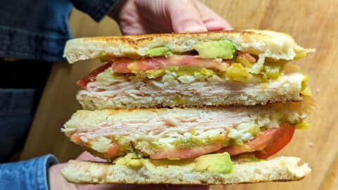 A person holds an Albuquerque Turkey Sandwich cut in half, revealing layers of sliced turkey, avocado, tomato, and possibly cheese on toasted bread, with a wooden surface in the background.