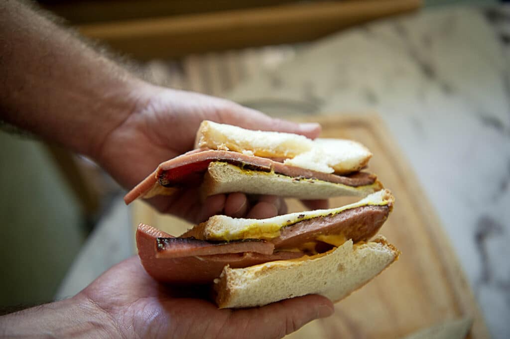 Close-up of two hands holding a Fried Bologna Sandwich cut in half, filled with slices of meat, cheese, and mustard on white sandwich bread. The background is slightly blurred, showing a wooden cutting board.