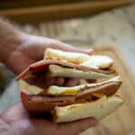 Close-up of two hands holding a Fried Bologna Sandwich cut in half, filled with slices of meat, cheese, and mustard on white sandwich bread. The background is slightly blurred, showing a wooden cutting board.