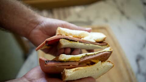 Close-up of two hands holding a Fried Bologna Sandwich cut in half, filled with slices of meat, cheese, and mustard on white sandwich bread. The background is slightly blurred, showing a wooden cutting board.