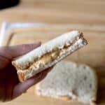 A hand holds half of a peanut butter sandwich made with white bread, its creamy filling visible—reminiscent of a classic Fluffernutter. Another sandwich half rests on a wooden surface in the background.