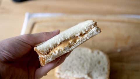 A hand holds half of a peanut butter sandwich made with white bread, its creamy filling visible—reminiscent of a classic Fluffernutter. Another sandwich half rests on a wooden surface in the background.