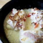 Three pieces of chicken breast in a cast iron skillet, inspired by the classic hot brown, topped with melted cheese, crispy bacon, and fresh tomato slices, with bubbling cheese around the edges.