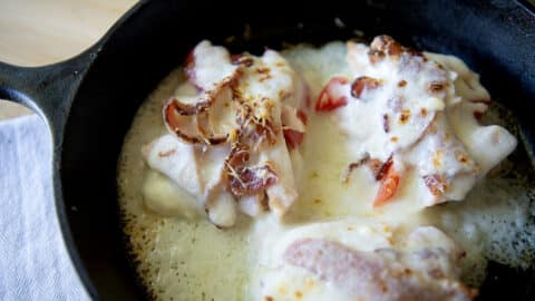 Three pieces of chicken breast in a cast iron skillet, inspired by the classic hot brown, topped with melted cheese, crispy bacon, and fresh tomato slices, with bubbling cheese around the edges.