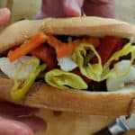 A hand holds an Italian Beef Sandwich filled with sliced beef, mozzarella cheese, pickled carrots, red peppers, and yellow pepper rings on a sub roll.
