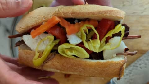 A hand holds an Italian Beef Sandwich filled with sliced beef, mozzarella cheese, pickled carrots, red peppers, and yellow pepper rings on a sub roll.