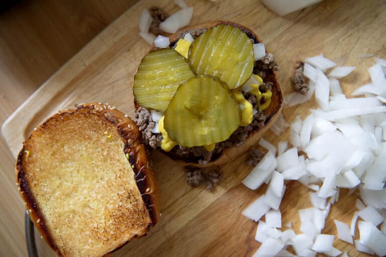 A Loose Meat Sandwich with toasted bread, ground beef, chopped onions, mustard, mayonnaise, and pickles sits on a wooden board beside a pile of diced raw onions.