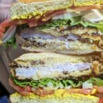 A close-up of a Pork Chop Sandwich with crispy fried chicken, lettuce, tomato, onions, and mustard on a bun. The sandwich is being held, showing its delicious layers clearly.