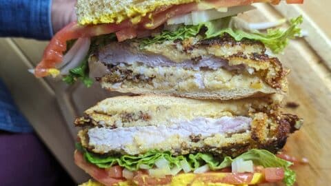 A close-up of a Pork Chop Sandwich with crispy fried chicken, lettuce, tomato, onions, and mustard on a bun. The sandwich is being held, showing its delicious layers clearly.
