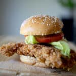 A crispy Fried Pork Tenderloin Sandwich with lettuce, tomato, and a sesame seed bun, placed on a wooden board.