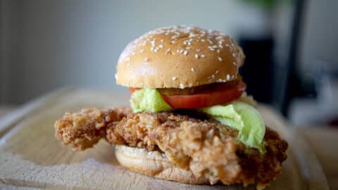 A crispy Fried Pork Tenderloin Sandwich with lettuce, tomato, and a sesame seed bun, placed on a wooden board.