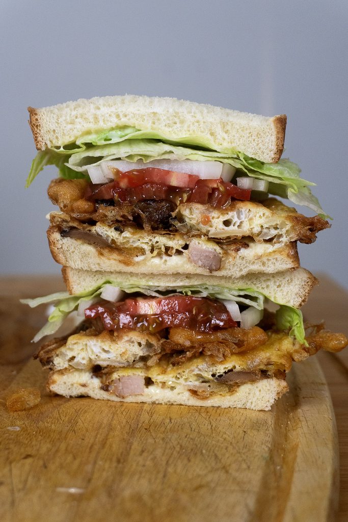 A St. Paul Sandwich cut in half, stacked with layers of lettuce, tomato, onion, bacon, and omelet between slices of white bread, sits invitingly on a wooden surface.