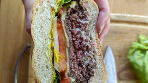 A hand holds half of a steamed burger with lettuce, tomato, melted cheese, a partially cooked beef patty, and mustard on a bun. A knife and lettuce leaves are on a wooden cutting board in the background.