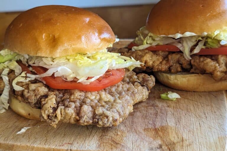 Two crispy chicken fried steak sandwiches with shredded lettuce, tomato slices, and soft golden buns are placed on a wooden cutting board.