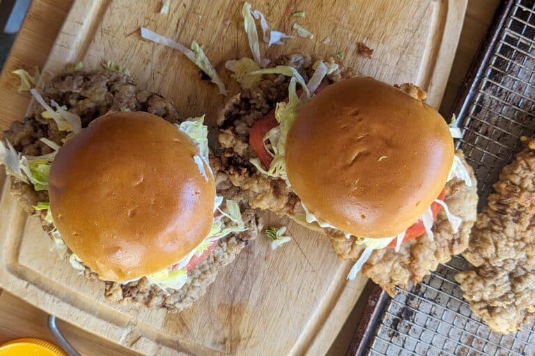 Two crispy chicken fried steak sandwiches with lettuce and tomato on shiny brioche buns, placed on a wooden cutting board next to a wire rack with more fried chicken.