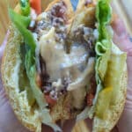 A person holds a Juicy Lucy Burger cut in half, revealing melted cheese, lettuce, pickles, tomato, and ground beef between two golden buns. The filling is juicy and layered with fresh vegetables.