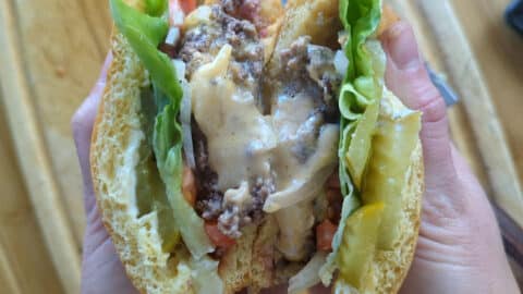 A person holds a Juicy Lucy Burger cut in half, revealing melted cheese, lettuce, pickles, tomato, and ground beef between two golden buns. The filling is juicy and layered with fresh vegetables.