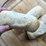 A hand holds a baked, golden-brown pepperoni roll with a slightly crisp crust. Two more similar pepperoni rolls rest on a wooden cutting board in the background.