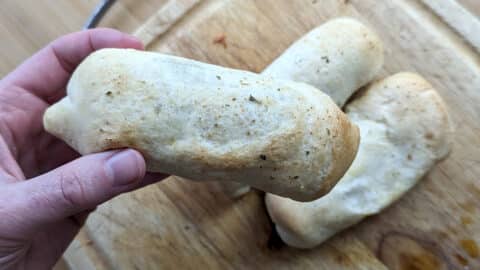 A hand holds a baked, golden-brown pepperoni roll with a slightly crisp crust. Two more similar pepperoni rolls rest on a wooden cutting board in the background.