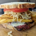 A Primanti Brothers Sandwich on thick white bread is filled with tomato slices, coleslaw, French fries, melted cheese, and grilled meat, all stacked high on a wooden cutting board.