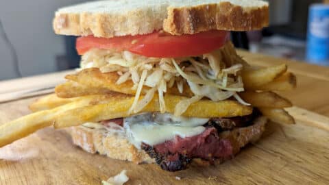 A Primanti Brothers Sandwich on thick white bread is filled with tomato slices, coleslaw, French fries, melted cheese, and grilled meat, all stacked high on a wooden cutting board.
