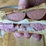 A hand holds a hearty Sailor Sandwich with two thick slices of sausage, layers of corned beef, melted cheese, and mustard between toasted bread. A knife and wooden cutting board are visible in the background.
