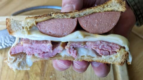 A hand holds a hearty Sailor Sandwich with two thick slices of sausage, layers of corned beef, melted cheese, and mustard between toasted bread. A knife and wooden cutting board are visible in the background.