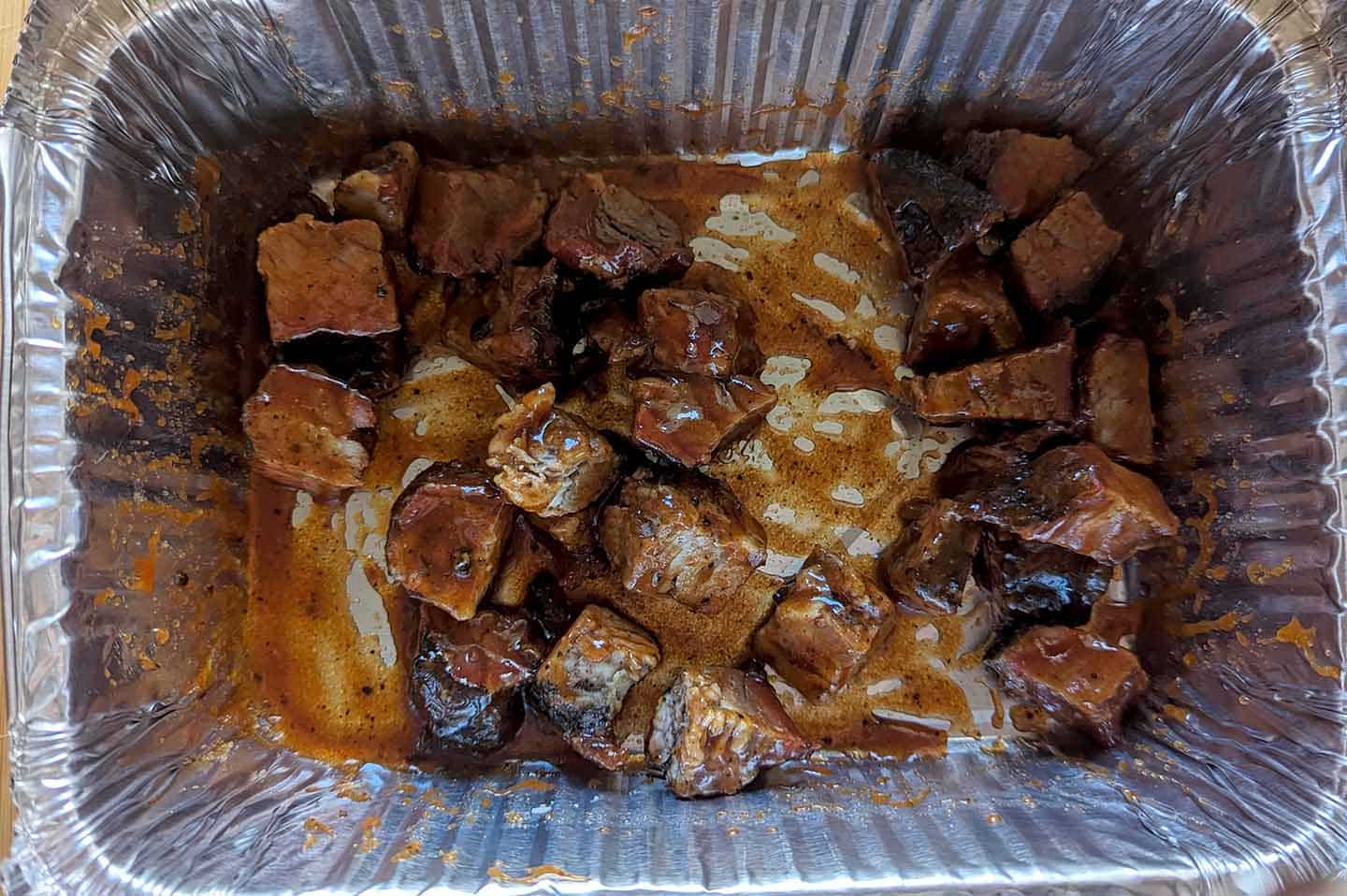 Chunks of cooked beef brisket coated in sauce sit in a disposable aluminum tray, with juices and bits of seasoning visible around the meat—perfect for piling onto a burnt ends sandwich.