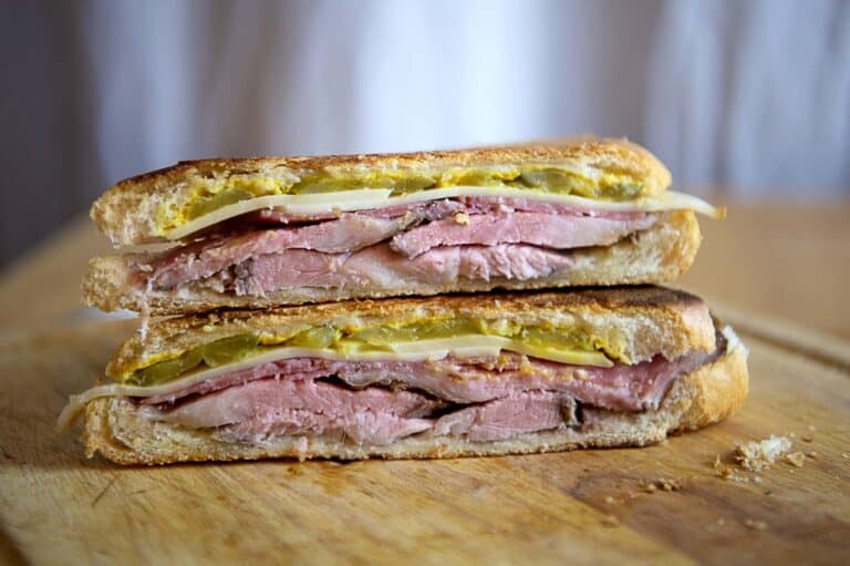 A classic Cubano sandwich cut in half, showing layers of ham, roast pork, Swiss cheese, pickles, and mustard inside toasted, pressed bread, resting on a wooden cutting board.