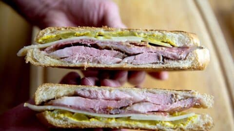 A close-up of two hands holding a halved Cubano, showing layers of roast pork, ham, Swiss cheese, pickles, and mustard between toasted bread.