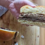 A hand holds half of a livermush sandwich with two beef patties and a bun, showing the cross-section. The other sandwich half and a large knife rest on a wooden cutting board in the background.