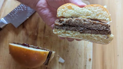 A hand holds half of a livermush sandwich with two beef patties and a bun, showing the cross-section. The other sandwich half and a large knife rest on a wooden cutting board in the background.
