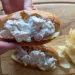 Two hands hold a classic lobster roll packed with creamy lobster salad over a wooden cutting board, with a small pile of potato chips on the side.