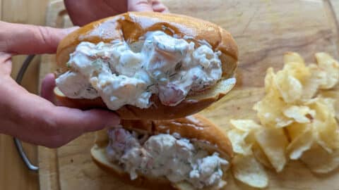 Two hands hold a classic lobster roll packed with creamy lobster salad over a wooden cutting board, with a small pile of potato chips on the side.