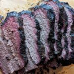 Sliced smoked brisket with a dark, charred bark and a pink smoke ring on a wooden cutting board, perfect for building a smoked brisket sandwich. The meat appears juicy and tender with visible seasoning on the surface.
