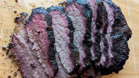 Sliced smoked brisket with a dark, charred bark and a pink smoke ring on a wooden cutting board, perfect for building a smoked brisket sandwich. The meat appears juicy and tender with visible seasoning on the surface.