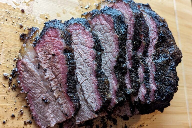 Sliced smoked brisket with a dark, charred bark and a pink smoke ring on a wooden cutting board, perfect for building a smoked brisket sandwich. The meat appears juicy and tender with visible seasoning on the surface.