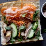 A partially open banh mi sandwich on a dark surface, filled with chicken, sliced cucumber, jalapeños, cilantro, pickled carrots and daikon, with a small bowl of pate on the side completes this classic banh mi spread.