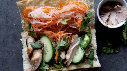 A partially open banh mi sandwich on a dark surface, filled with chicken, sliced cucumber, jalapeños, cilantro, pickled carrots and daikon, with a small bowl of pate on the side completes this classic banh mi spread.