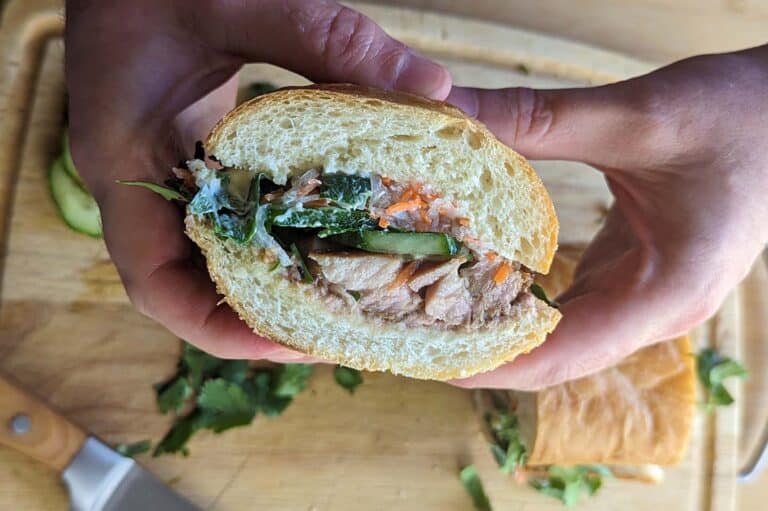A person holds a banh mi sandwich with shredded meat, cucumbers, greens, and pickled vegetables in a crusty baguette, above a wooden cutting board with a knife and scattered herbs.