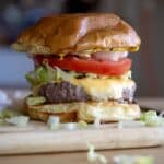 A juicy bison burger with lettuce, tomato, onion, and melted cheese in a brioche bun sits on a wooden board, with pieces of chopped lettuce and onion scattered around.