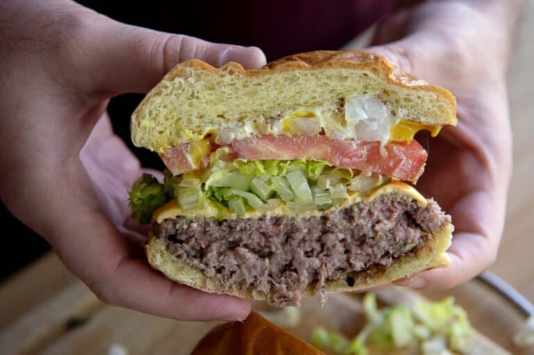 A close-up of hands holding a hamburger cut in half, showing layers of lettuce, tomato, pickles, onions, cheese, and a beef patty inside a soft bun.